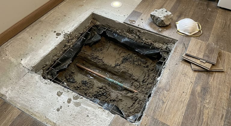 Square opening broken through a concrete floor slab showing the slab edge in section, a vapor barrier, damp sand fill and a stained copper water line lying in standing water below.