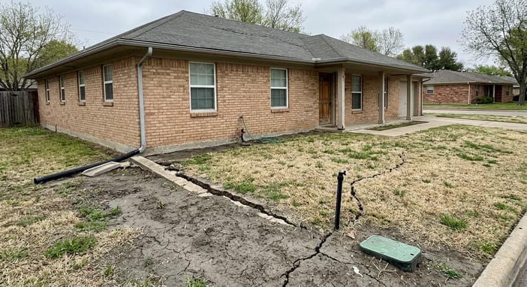 Front of a single-story brick ranch house on a concrete slab, with dried shrinkage cracks running through dark clay soil in the lawn up to the slab edge.