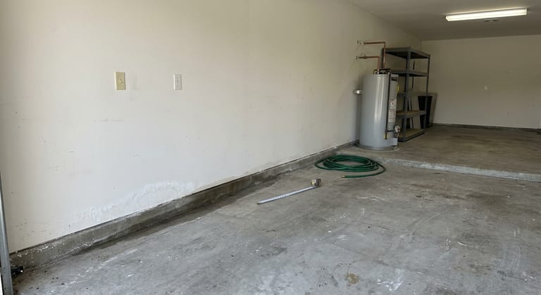 Dark damp strip along the base of a garage wall on a concrete slab, with white mineral bloom low on the wall and a gas water heater standing on the same floor a few feet away.