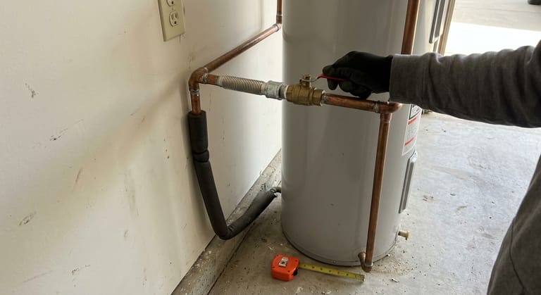 Gloved hand closing the brass ball valve on the cold inlet of a water heater standing in a garage, with copper lines above the tank and the hot line running down the wall into the concrete floor.